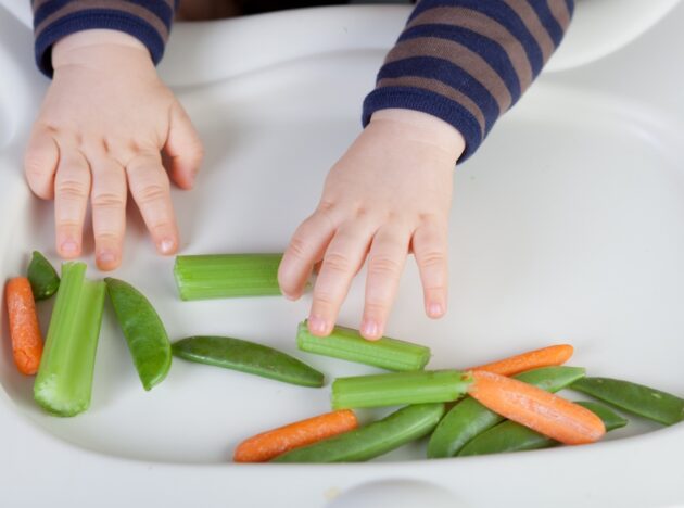 Baby reaching for loose vegetables on the front of their baby seat