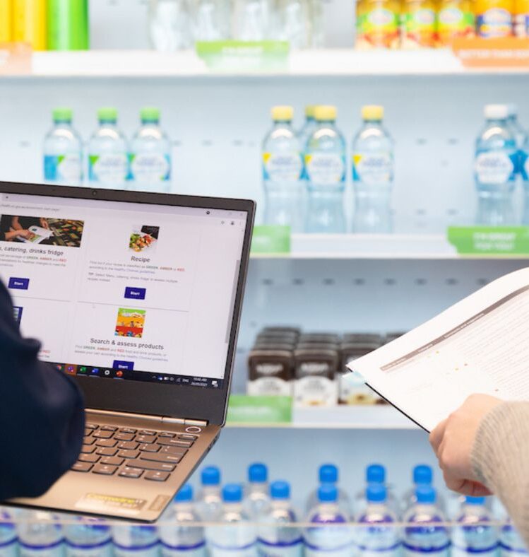 person using a laptop and another person holding a booklet in front of shelves of drinks