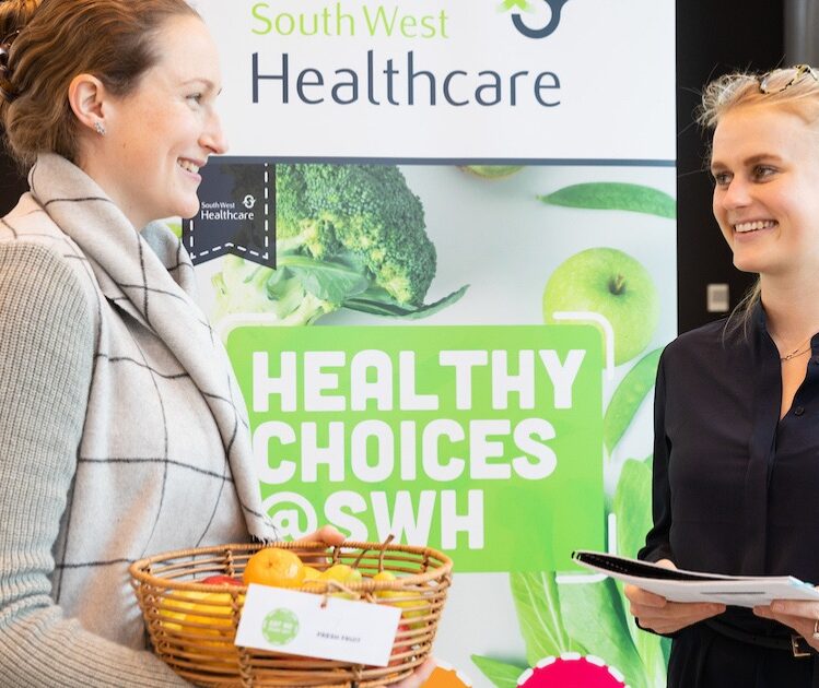 a woman holding a bowl of fruit talking to another woman holding a booklet