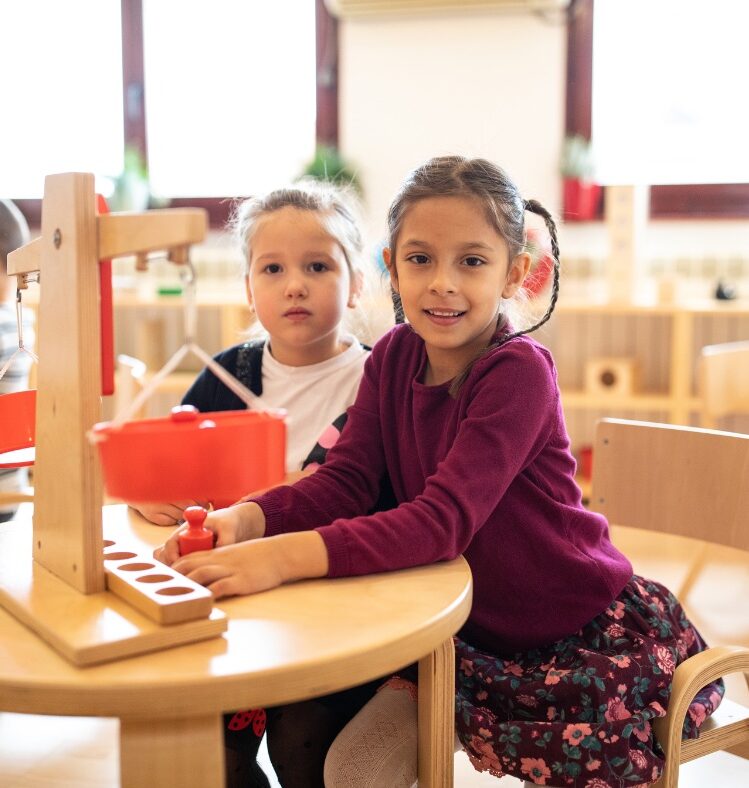 Two girls sitting at a table in a classroom with a toy scale
