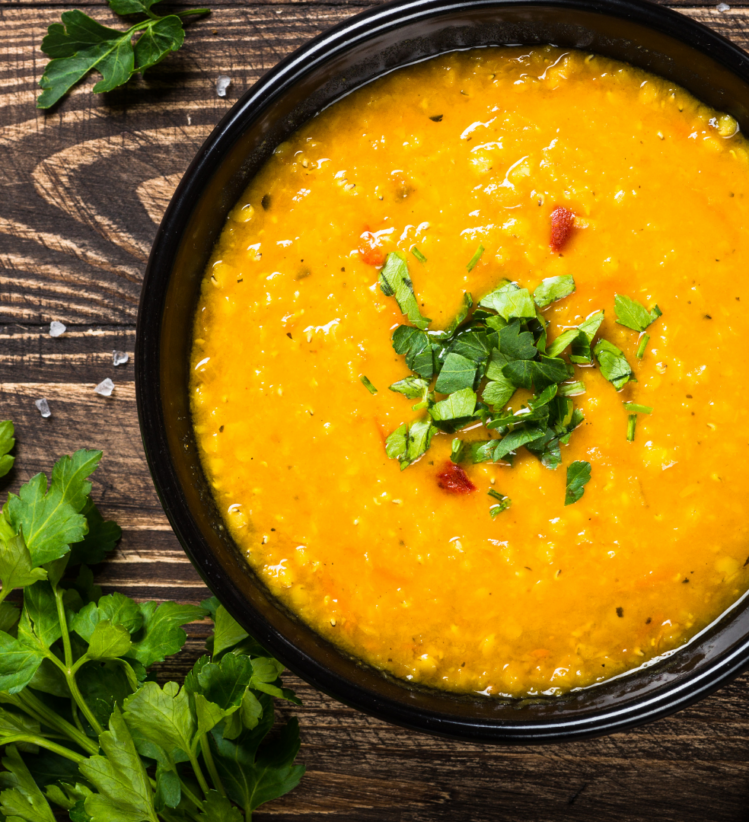 a bowl of carrot lentil soup next to a spoon and sprig of herbs