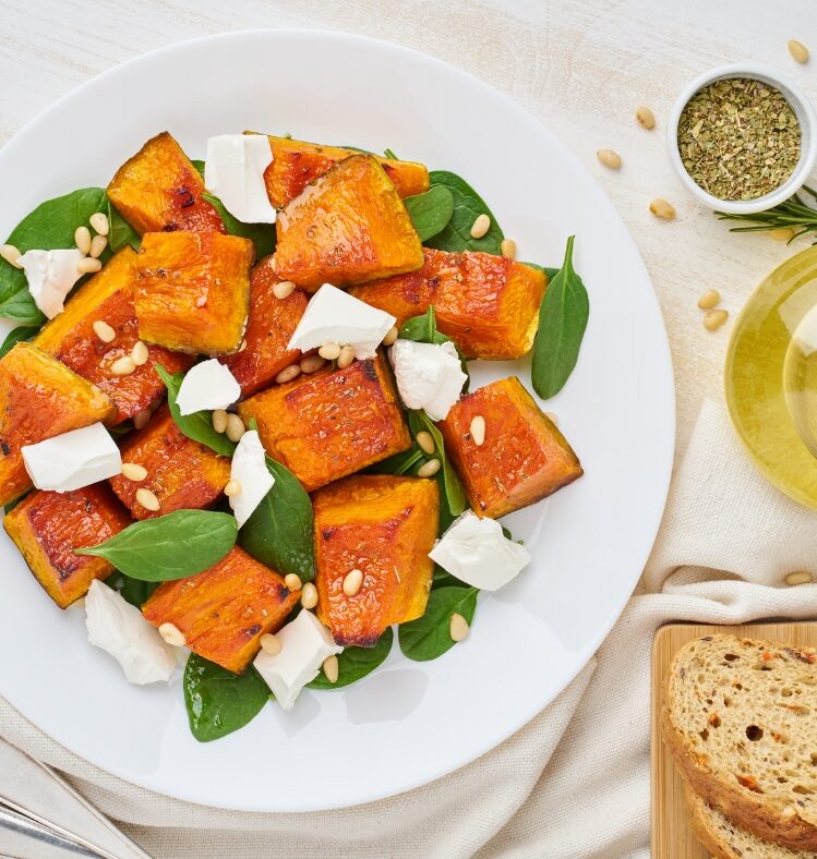 Roasted vegetable, spinach, pine nut and feta salad on na plate next to some sliced bread