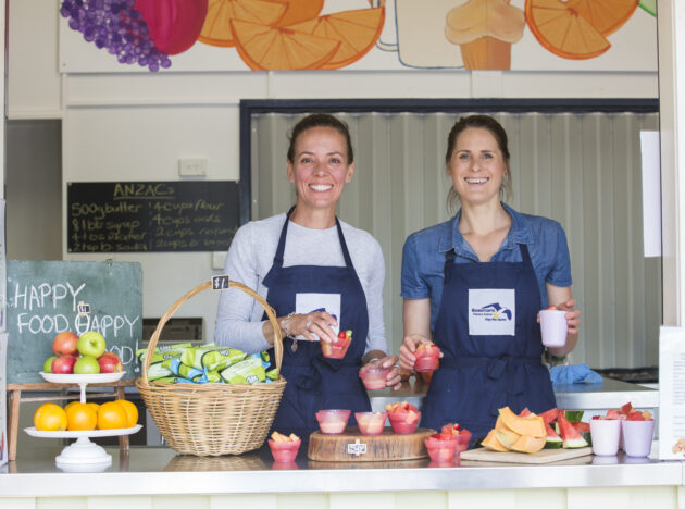 Two women working at the counter of a canteen and smiling while holding fruit cups